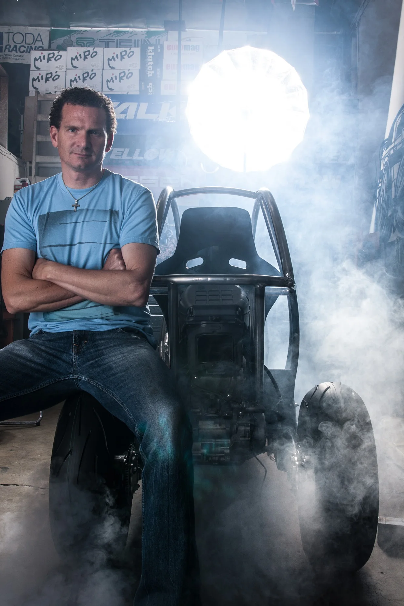 Markus Leo Scholten seated with arms crossed beside a bare tubular three-wheel prototype chassis in a backlit workshop